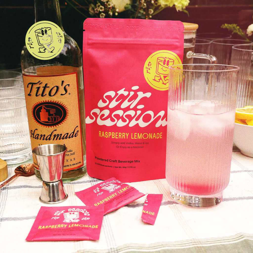 Bottle of Tito's Handmade Vodka and Stir Sessions Raspberry Lemonade powdered cocktail or mocktail drink beverage packaging, and a glass with pink lemonade on a table.