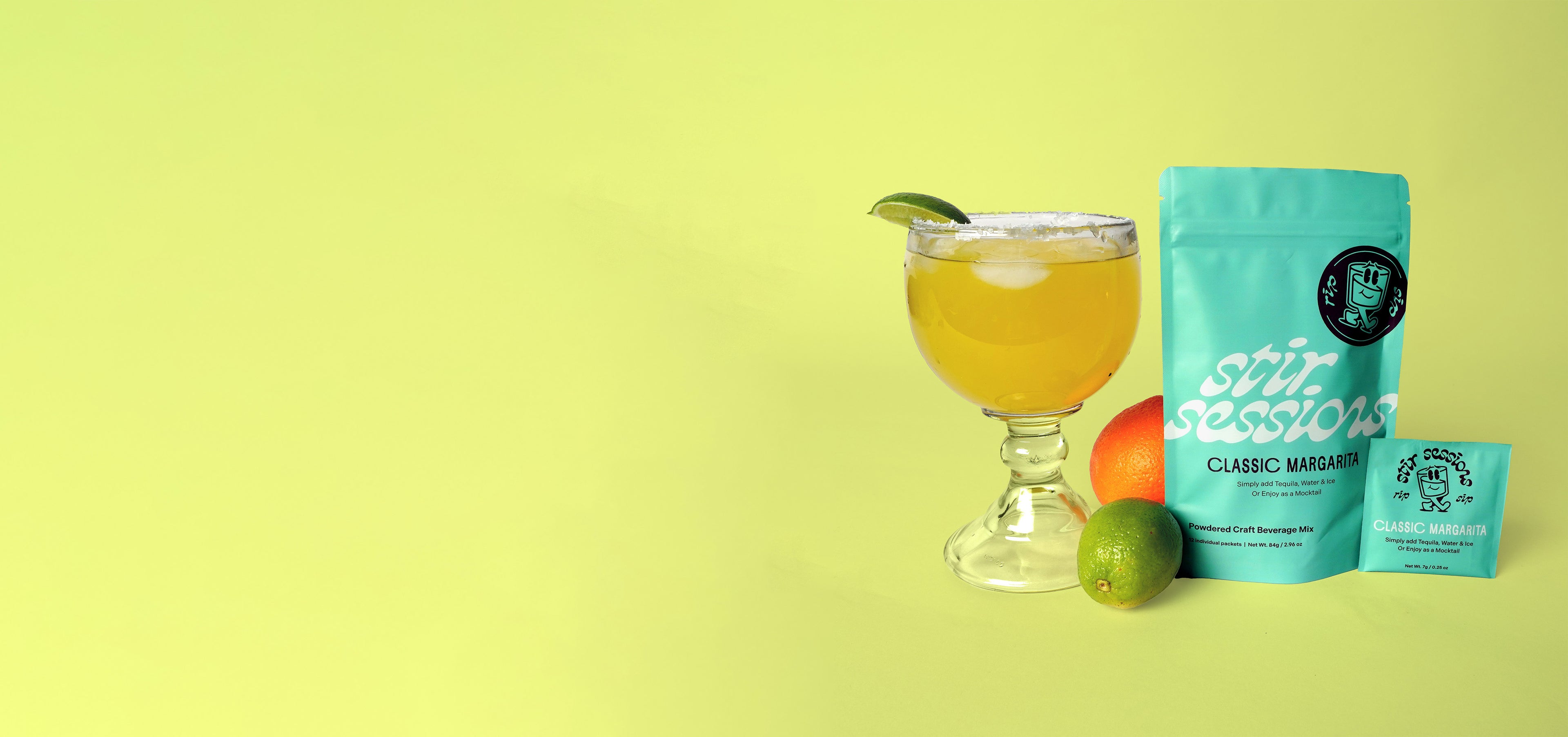 Cocktail glass with a drink, lime, and oranges next to a bag of 'Stir Sessions' Classic Margarita powdered cocktail or mocktail drink beverage mix on a yellow background