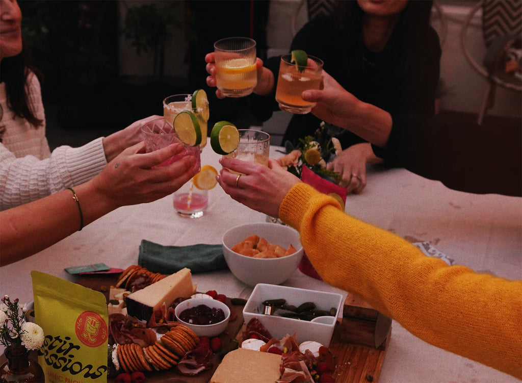 People toasting with Stir Sessions Cocktail drinks at a table with snacks and appetizers.