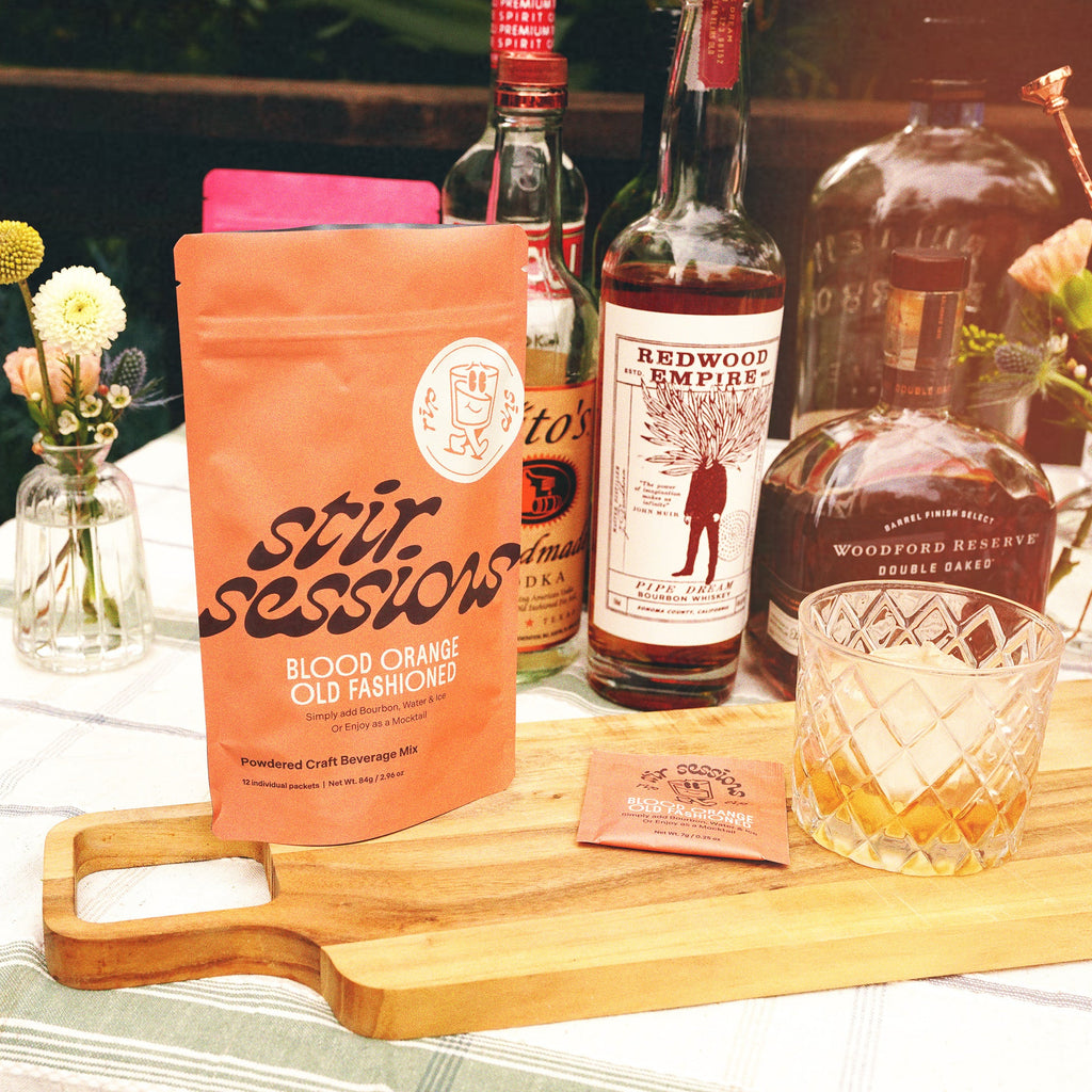 Stir Sessions Blood Orange Old Fashioned powdered cocktail or mocktail drink beverage mix packaging with bourbon bottles, and a glass on a table with a wooden cutting board.